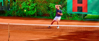 Eine Person spielt Tennis auf einem Sandplatz, umgeben von gr&uuml;ner Vegetation und einem Zaun mit Werbebannern.