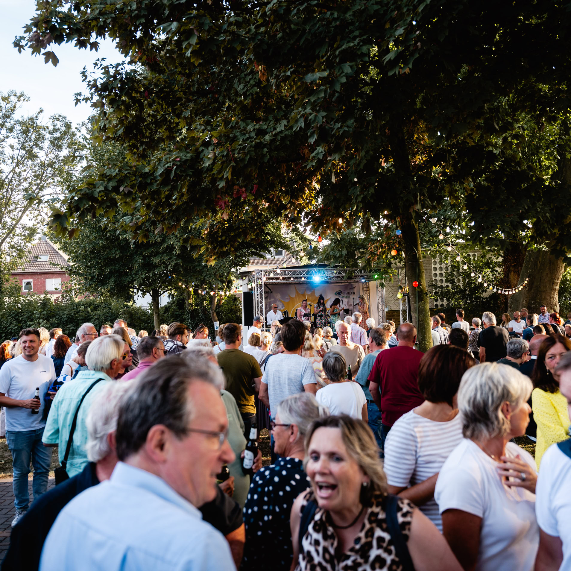 Eine Menschenmenge versammelt sich im Freien unter B&auml;umen, einige unterhalten sich, im Hintergrund sind H&auml;user zu sehen.