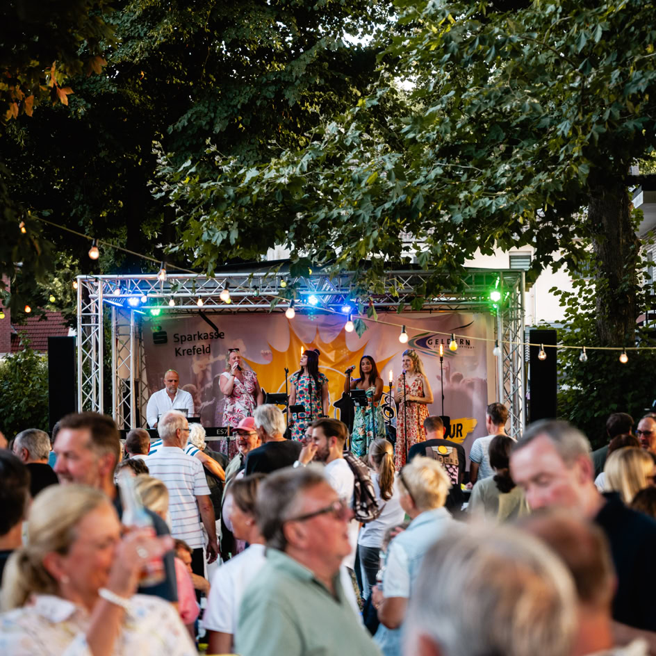 Eine Menschenmenge steht vor einer B&uuml;hne im Freien, auf der eine Band unter bunten Lichtern spielt, umgeben von B&auml;umen.