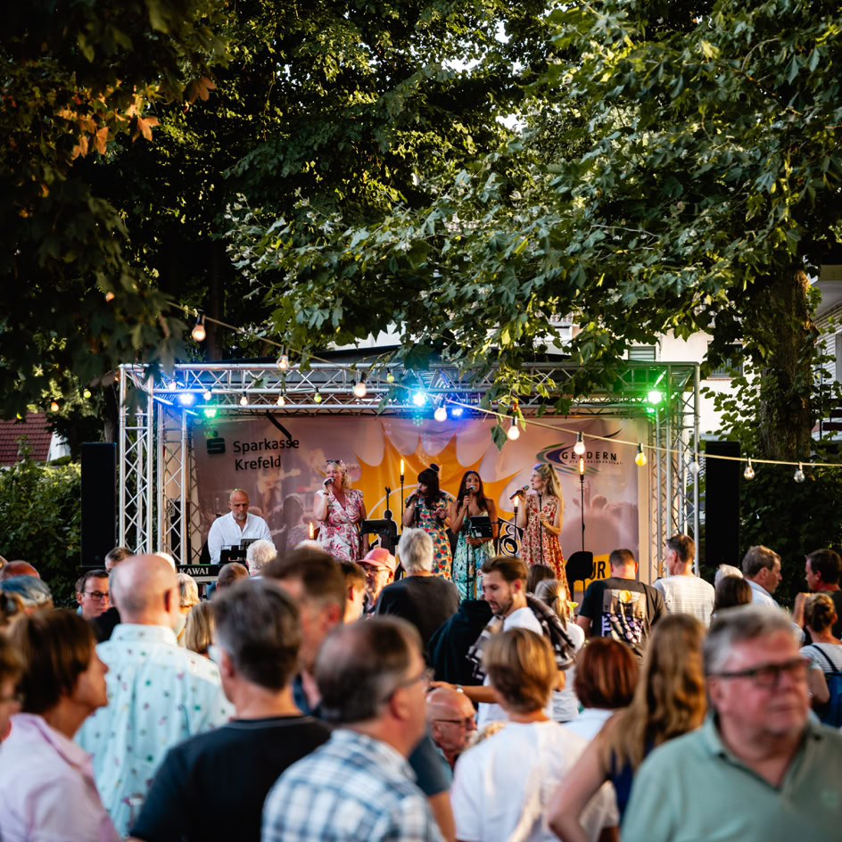 Eine Menschenmenge steht vor einer B&uuml;hne im Freien, auf der eine Band unter bunten Lichtern spielt, umgeben von B&auml;umen.