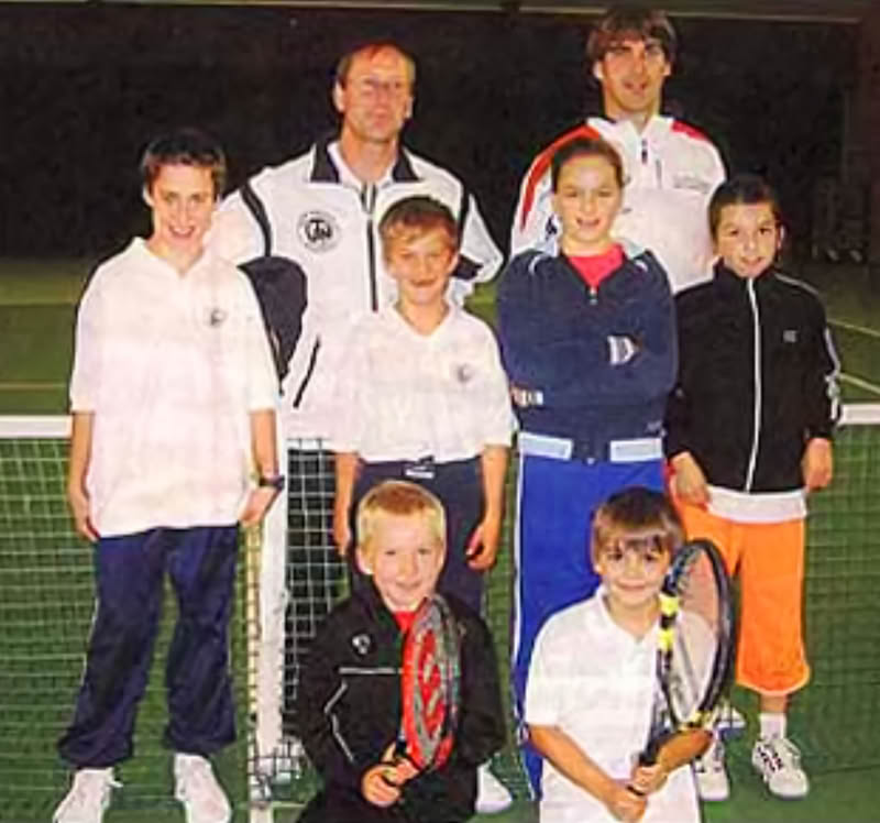 Eine Gruppe von sieben Personen, darunter zwei Erwachsene und f&uuml;nf Kinder, steht auf einem Tennisplatz vor einem Netz. Die Kinder halten Tennisschl&auml;ger.