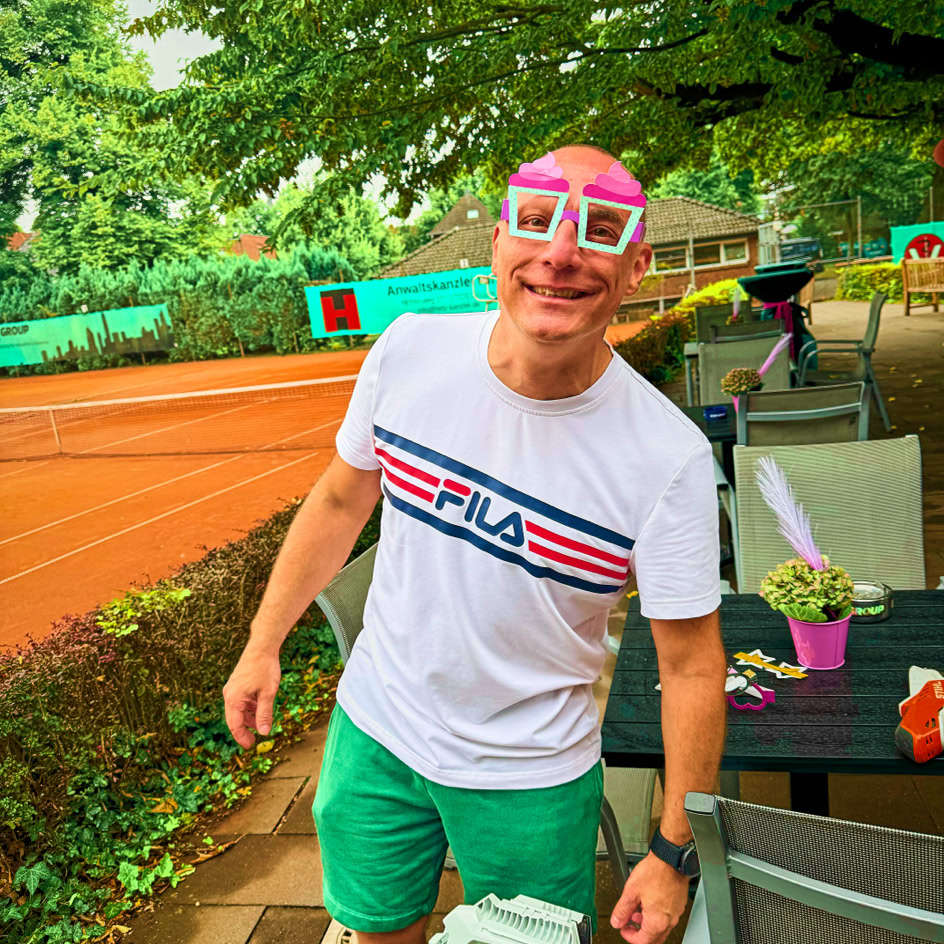 Ein Mann mit rosa Brille und wei&szlig;em T-Shirt steht l&auml;chelnd auf einem Tennisplatz neben einem Tisch mit Pflanzen.