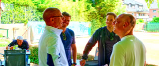 Mehrere Personen stehen drau&szlig;en unter einer &Uuml;berdachung und unterhalten sich. Im Hintergrund sind B&auml;ume und ein Geb&auml;ude sichtbar.