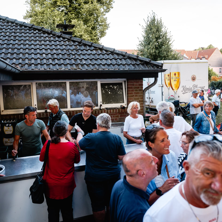 Menschen stehen an einem Tresen im Freien, einige unterhalten sich, w&auml;hrend andere Getr&auml;nke servieren. Im Hintergrund sind B&auml;ume und H&auml;user zu sehen.