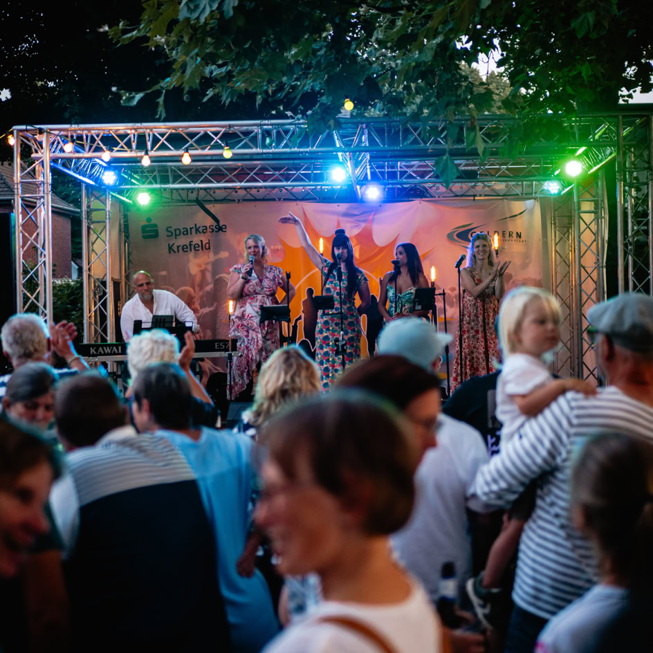 Eine Menschenmenge steht vor einer B&uuml;hne im Freien, auf der mehrere Personen unter bunten Lichtern auftreten.
