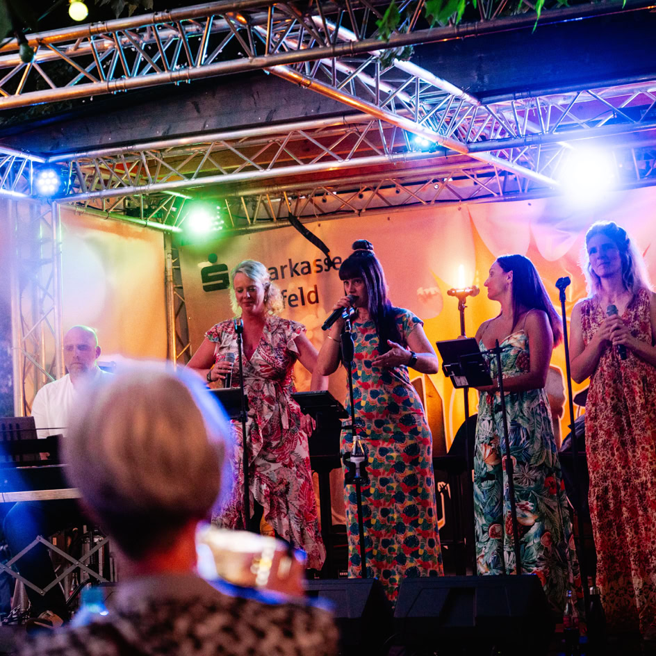 Eine Gruppe von Menschen steht auf einer B&uuml;hne unter bunten Lichtern, einige halten Mikrofone. Im Vordergrund ist eine Person mit grauen Haaren, die ein Smartphone h&auml;lt.