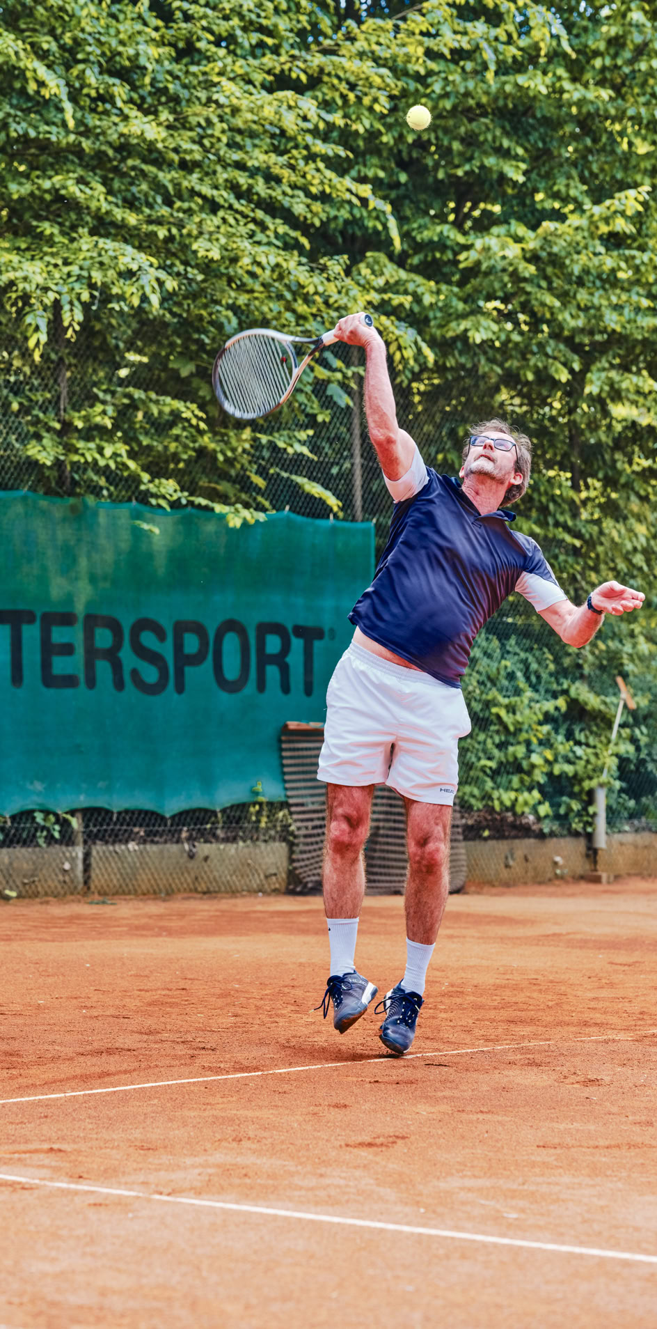 Ein Tennisspieler in blauer Kleidung und wei&szlig;en Shorts schl&auml;gt auf einem Sandplatz einen Ball auf, im Hintergrund sind gr&uuml;ne B&auml;ume und ein Werbebanner zu sehen.