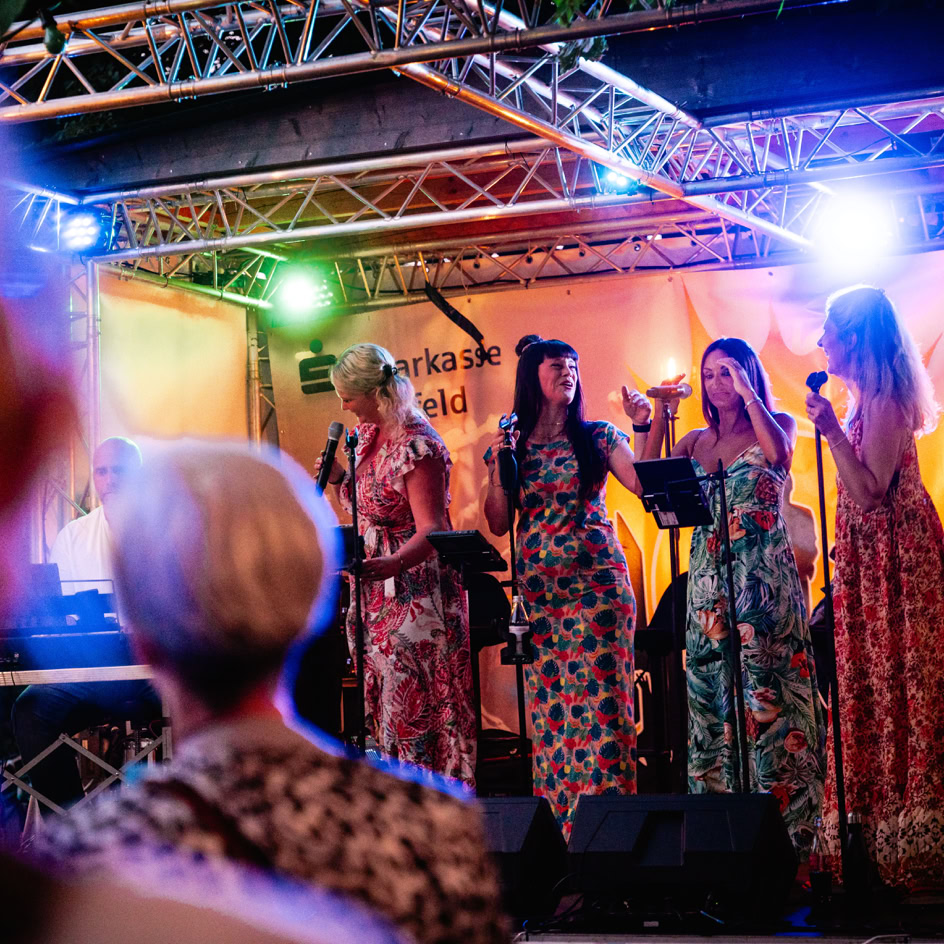 Vier Personen singen auf einer B&uuml;hne mit Mikrofonen, bunte Lichter im Hintergrund, Publikum im Vordergrund.