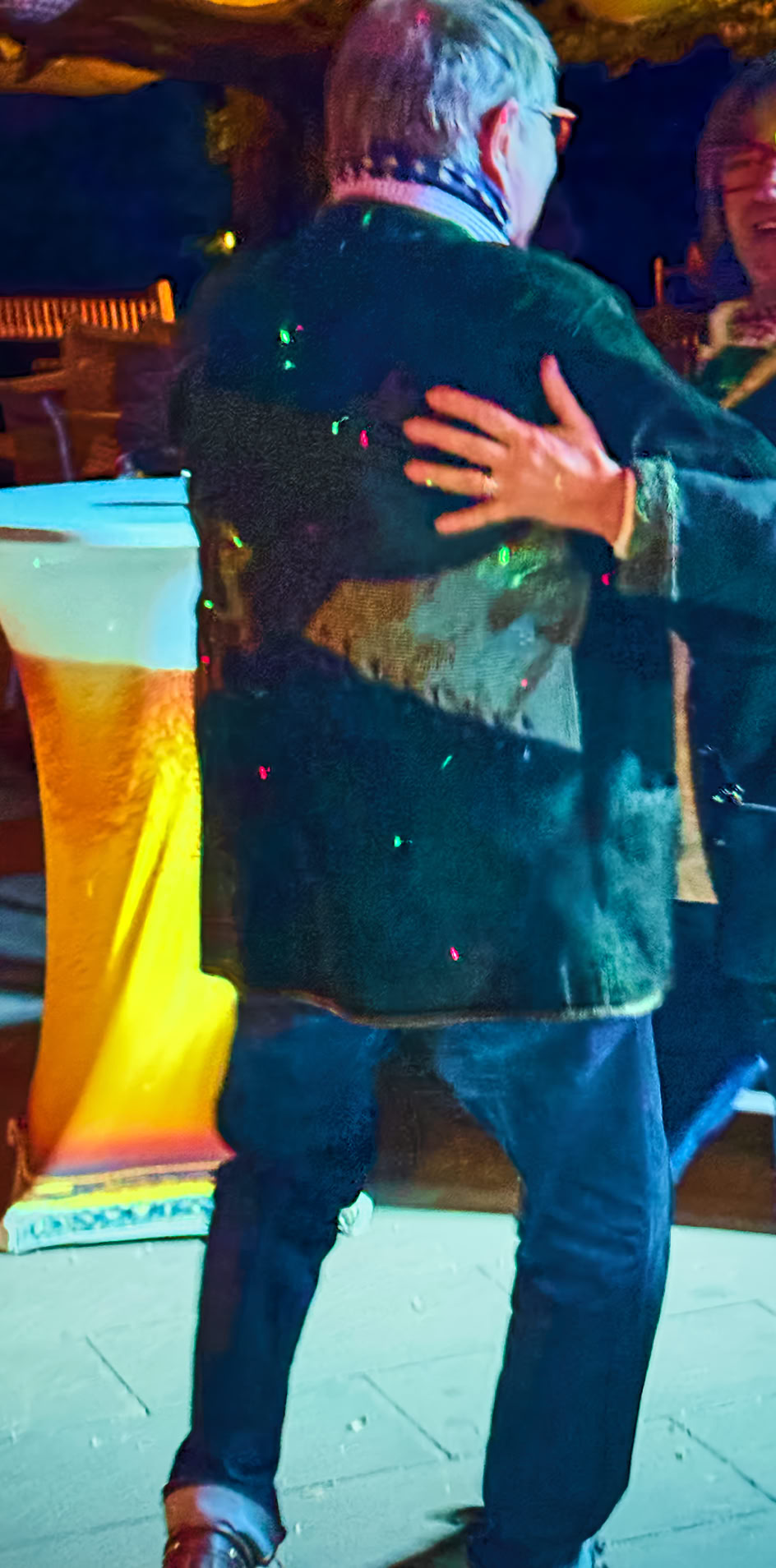 Zwei Personen tanzen zusammen in einem beleuchteten Raum, im Hintergrund steht ein Stehtisch.