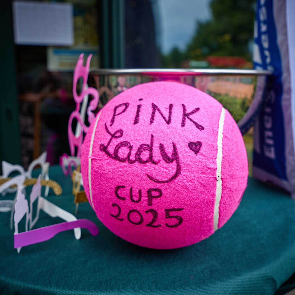 Ein pinker Tennisball mit der Aufschrift "Pink Lady Cup 2025" auf einem Tisch, daneben liegen pinke und wei&szlig;e Brillen.