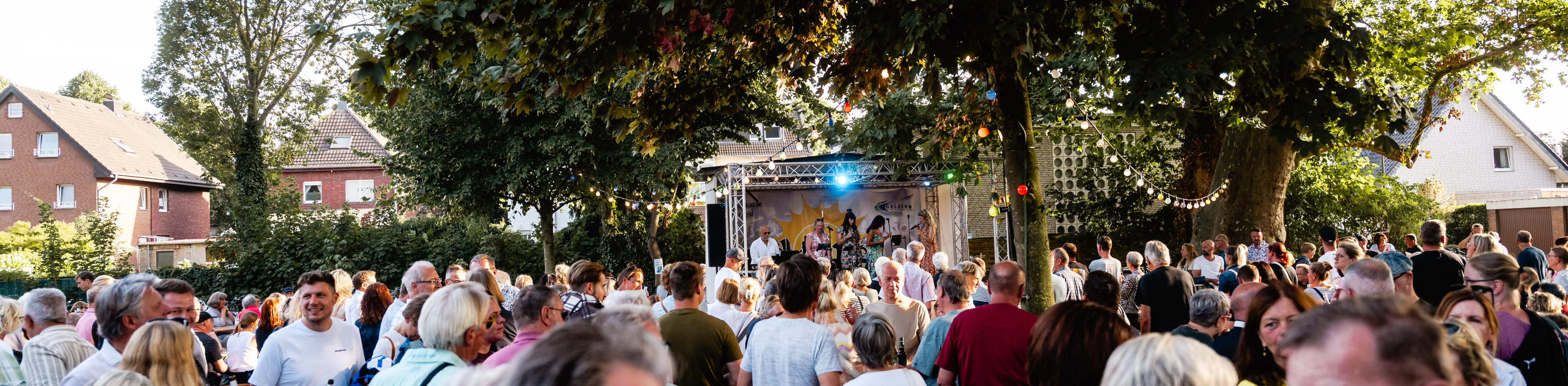 Eine Menschenmenge versammelt sich im Freien unter B&auml;umen, einige unterhalten sich, im Hintergrund sind H&auml;user zu sehen.