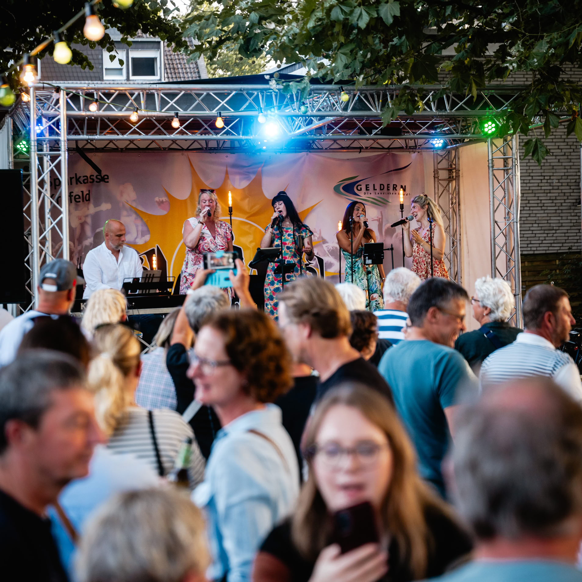 Menschenmenge vor einer B&uuml;hne im Freien mit Musikern, bunte Lichter und B&auml;ume im Hintergrund.