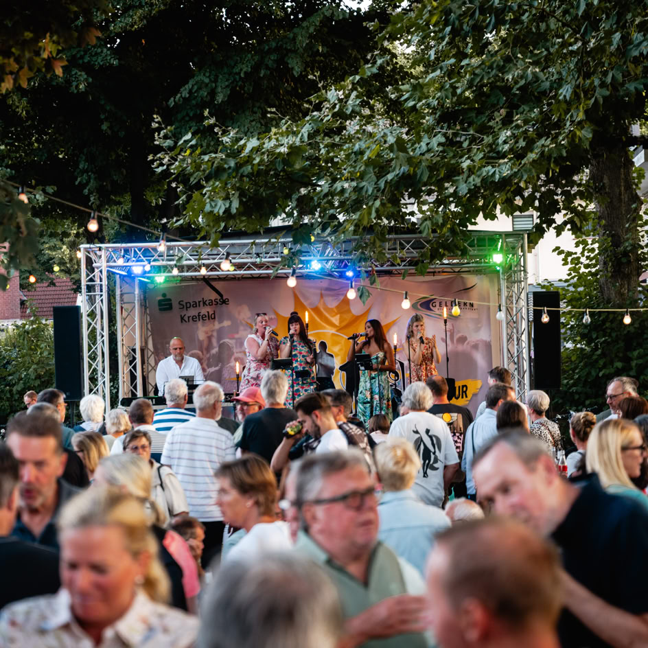 Eine Menschenmenge steht vor einer B&uuml;hne im Freien, auf der eine Band unter bunten Lichtern spielt; B&auml;ume und Lichterketten im Hintergrund.