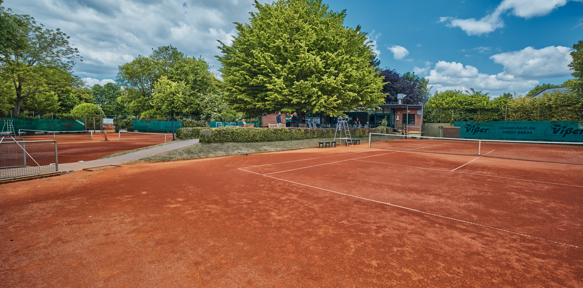 Ein Tennisplatz mit rotem Sandboden, umgeben von B&auml;umen und einem Zaun, unter einem blauen Himmel mit einigen Wolken.