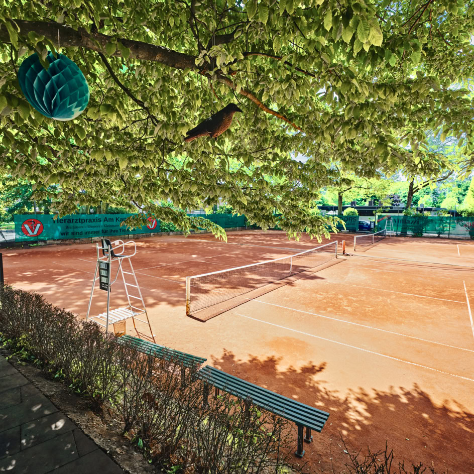 Tennisplatz mit rotem Sand, umgeben von B&auml;umen und gr&uuml;nen Netzen, im Vordergrund ein Baum mit h&auml;ngender Dekoration.