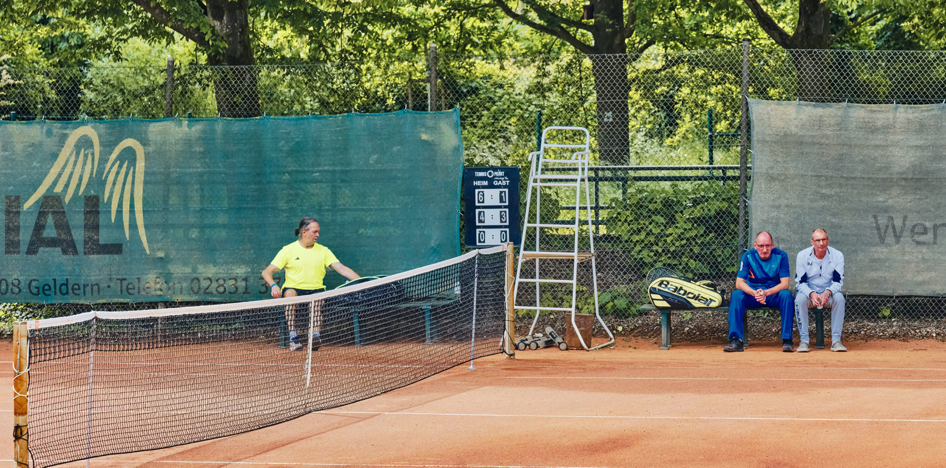 Ein Tennisplatz mit einem Spieler in gelbem Shirt, der den Ball schl&auml;gt, und zwei Personen, die auf einer Bank am Rand sitzen. Im Hintergrund sind B&auml;ume und ein Zaun zu sehen.