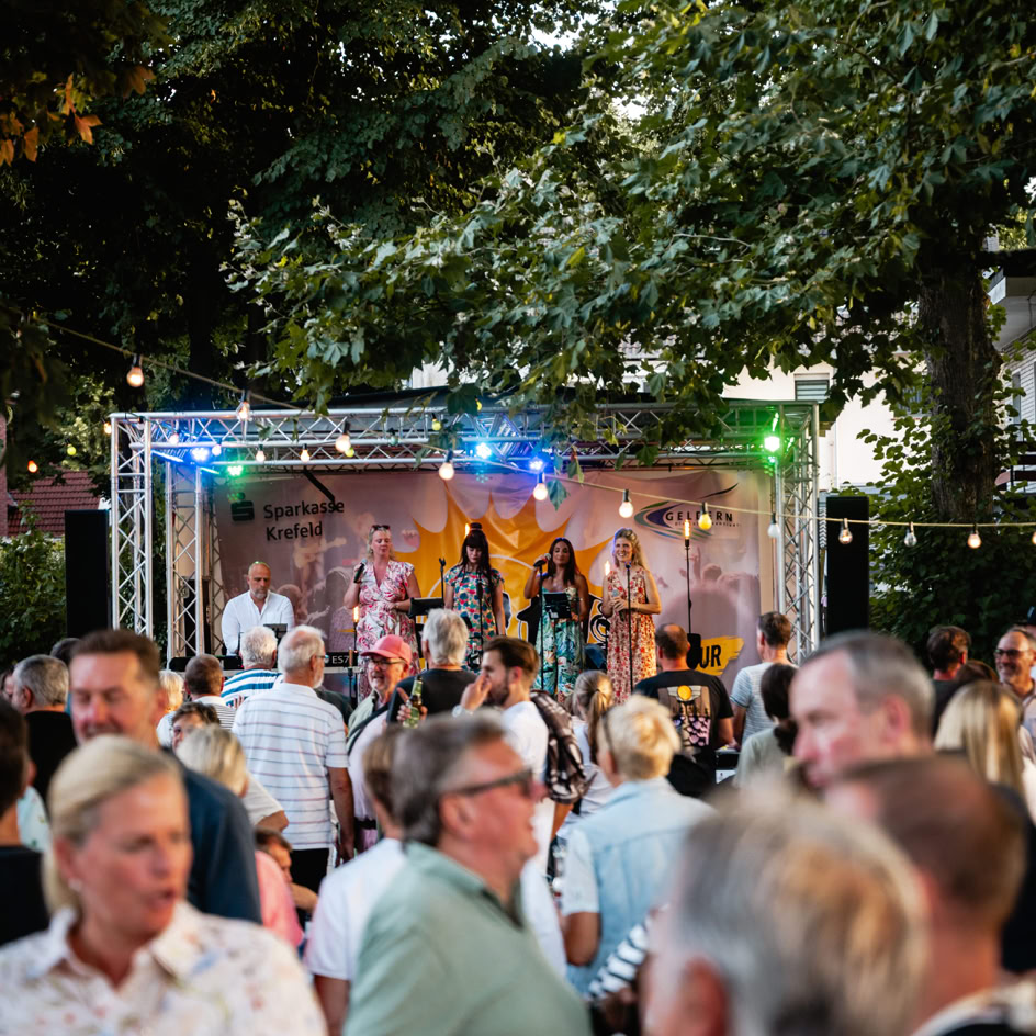 Menschenmenge vor einer B&uuml;hne im Freien, auf der eine Band unter bunten Lichtern spielt; umgeben von B&auml;umen.