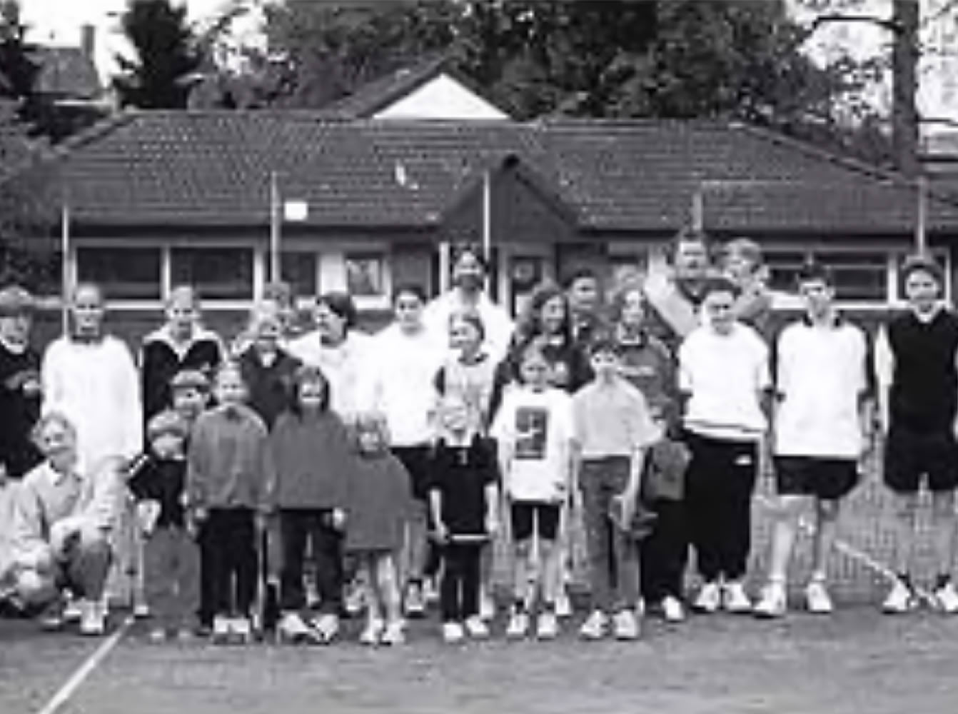 Gruppe von Menschen auf einem Tennisplatz, einige stehen, andere hocken; im Hintergrund sind B&auml;ume und Geb&auml;ude zu sehen.
