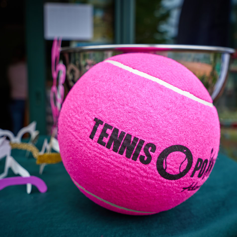 Ein gro&szlig;er, pinker Tennisball mit der Aufschrift "TENNIS" liegt auf einem gr&uuml;nen Tisch, im Hintergrund ist eine silberne Schale zu sehen.