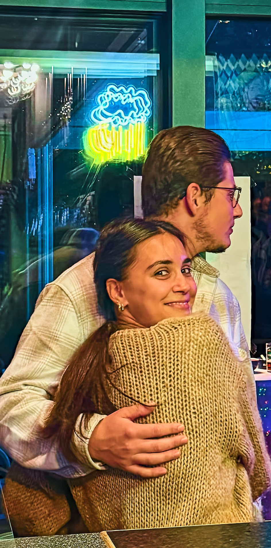 Ein Mann und eine Frau umarmen sich in einem Raum mit blauer Beleuchtung, im Hintergrund sind Blumen und eine Glast&uuml;r zu sehen.