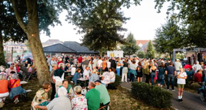 Eine gro&szlig;e Menschenmenge versammelt sich im Freien zwischen B&auml;umen, einige sitzen an Tischen, w&auml;hrend andere stehen.