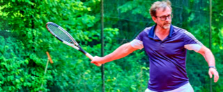 Ein Mann in einem lila Hemd spielt Tennis auf einem Platz im Freien, umgeben von gr&uuml;ner Vegetation.