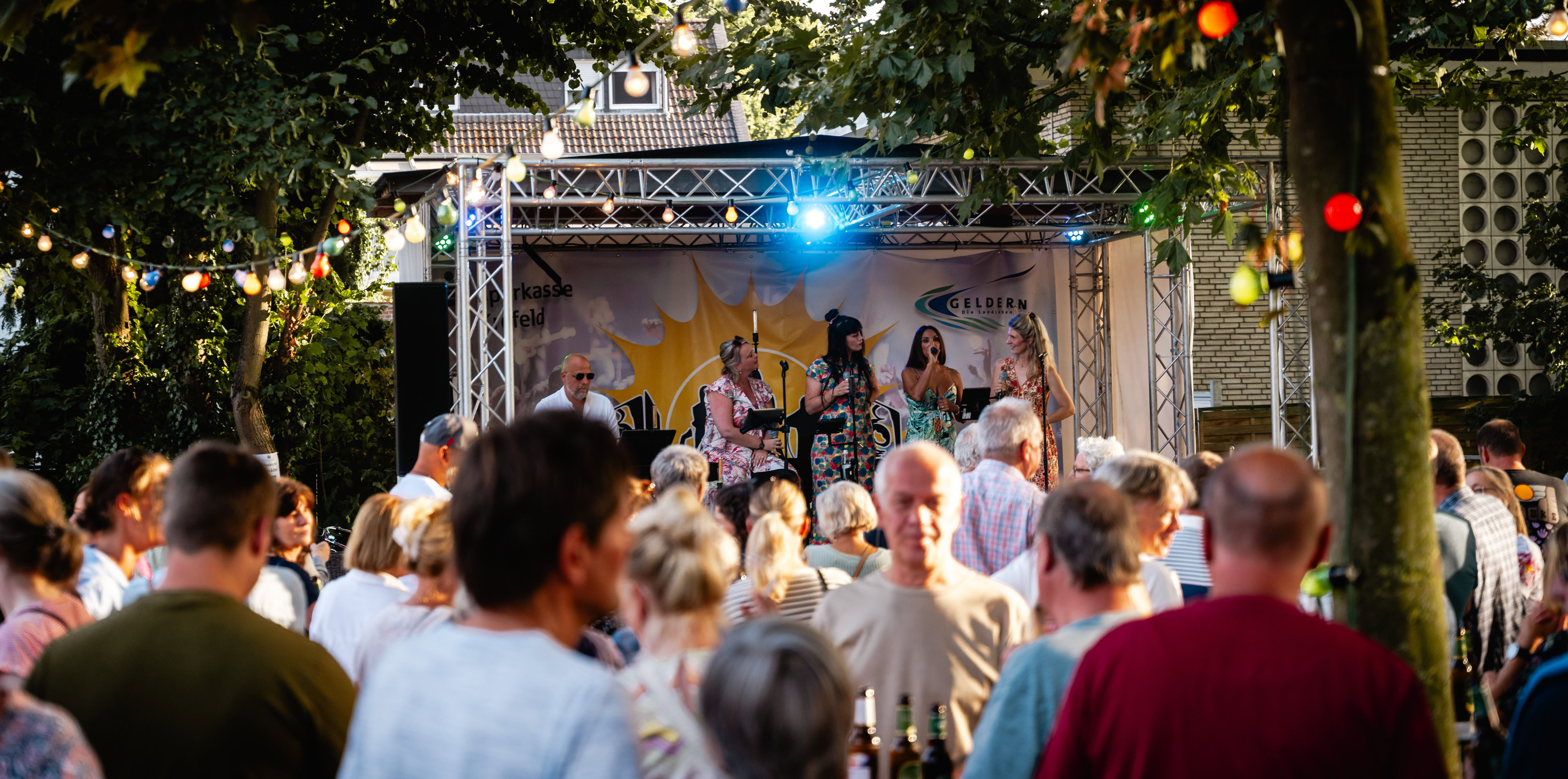 Eine Menschenmenge steht vor einer B&uuml;hne im Freien, auf der eine Band spielt; bunte Lichter h&auml;ngen in den B&auml;umen.