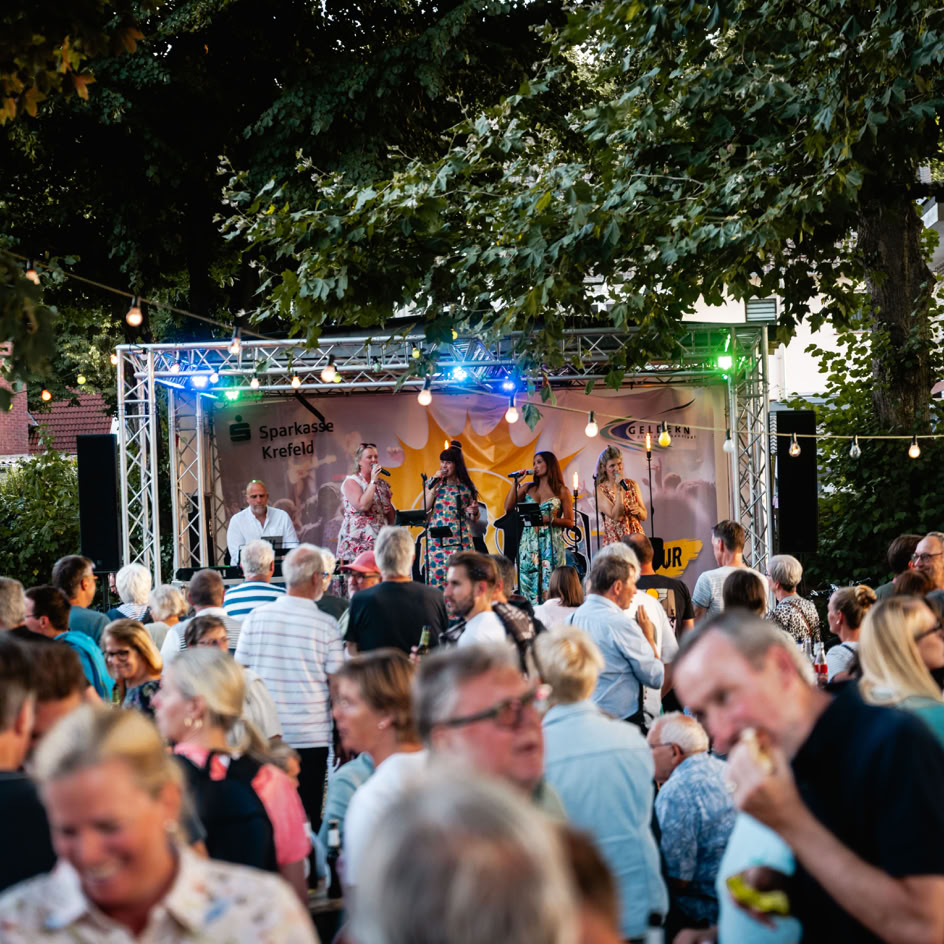 Eine Menschenmenge steht vor einer B&uuml;hne im Freien, auf der eine Band unter Lichtern spielt; umgeben von B&auml;umen und beleuchteten Girlanden.