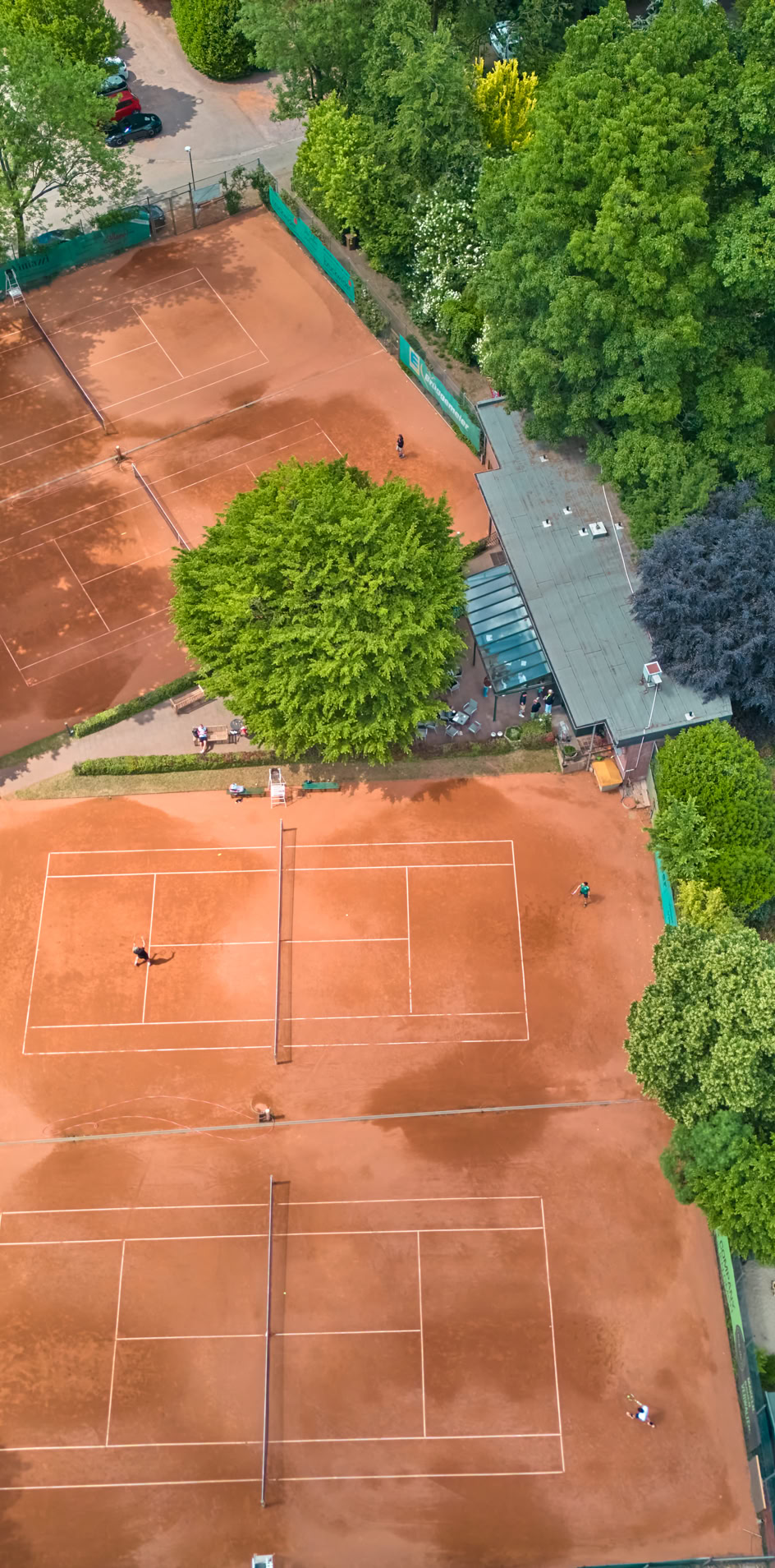 Luftaufnahme von zwei Sandtennispl&auml;tzen, umgeben von B&auml;umen und Geb&auml;uden.