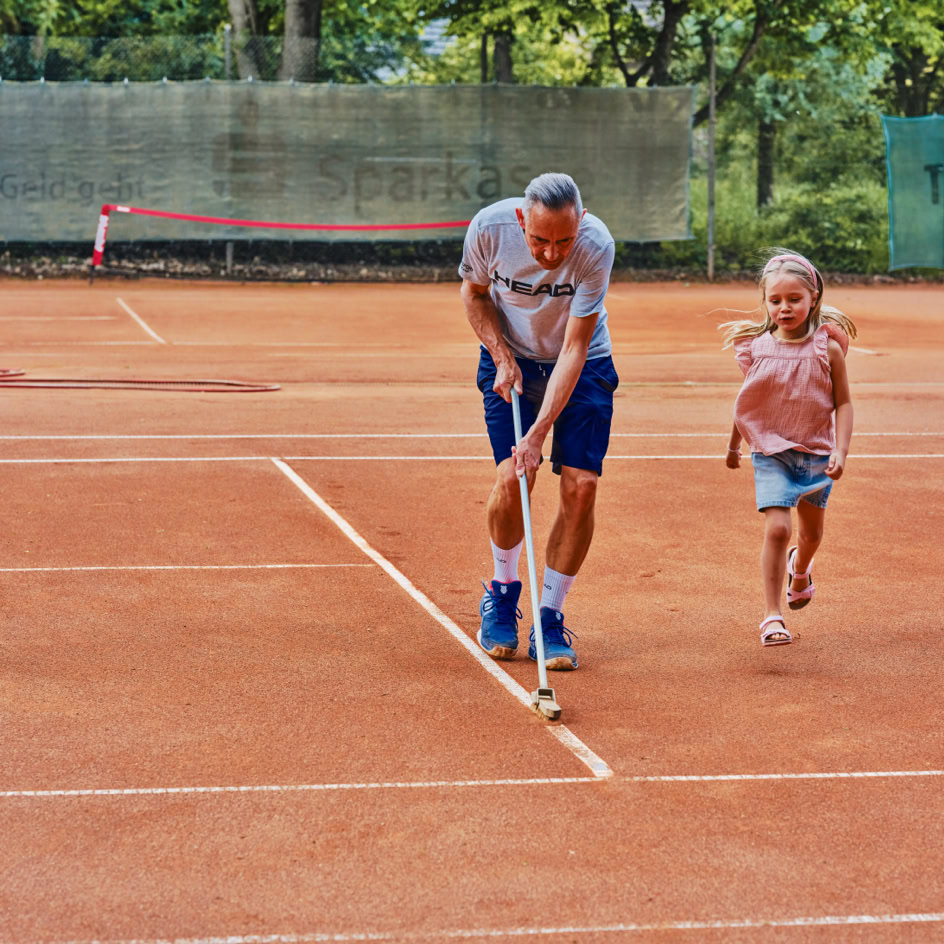 Ein Mann und ein Kind ziehen eine Linie auf einem Sandtennisplatz.