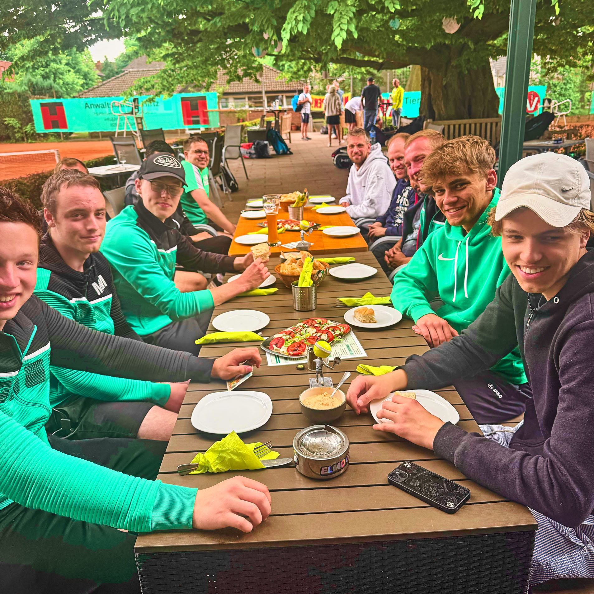 Eine Gruppe von Personen sitzt an einem langen Tisch im Freien, mit Tellern und Getr&auml;nken vor sich. Einige tragen gr&uuml;ne Jacken. Im Hintergrund sind B&auml;ume und andere Sitzbereiche zu sehen.