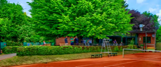 Ein Tennisplatz mit rotem Sand, umgeben von gr&uuml;nen B&auml;umen und einem Backsteingeb&auml;ude im Hintergrund.