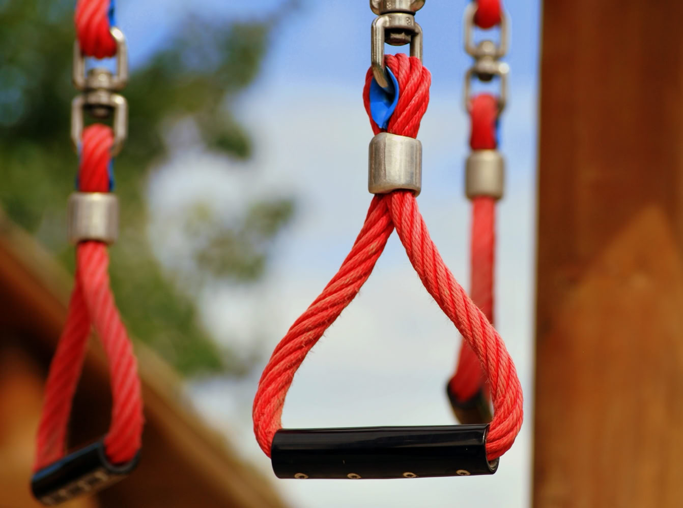 Rote Seilgriffe mit Metallbefestigungen an einem Spielplatzger&auml;t, unscharfer Hintergrund mit B&auml;umen und Himmel.