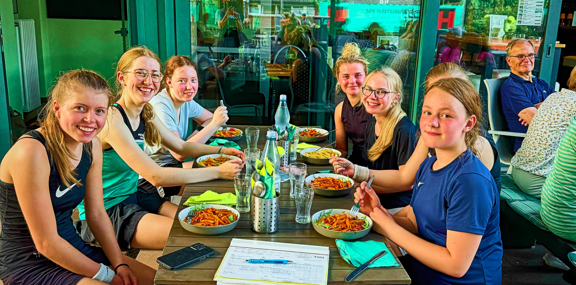 Sechs Personen sitzen an einem Tisch im Freien und essen, mit Getr&auml;nken und Tellern vor sich. Im Vordergrund stehen zwei Rucks&auml;cke.