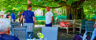 Mehrere Personen sitzen und stehen in einem Gartenbereich mit Tischen und St&uuml;hlen unter einem gro&szlig;en Baum.