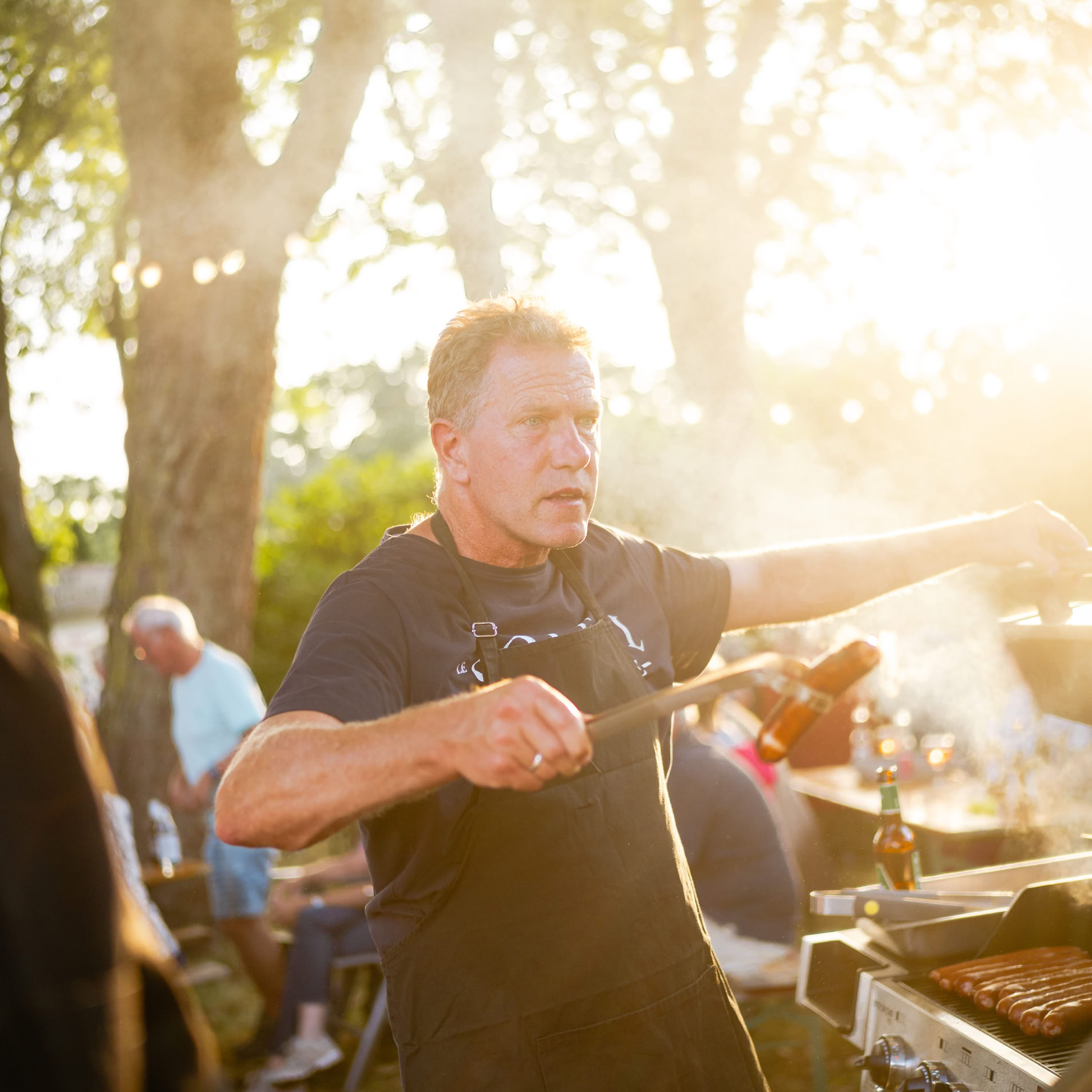Mann grillt im Freien bei Sonnenuntergang, umgeben von B&auml;umen und anderen Personen im Hintergrund.