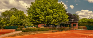 Ein Tennisplatz mit rotem Sand, umgeben von B&auml;umen und einem Backsteingeb&auml;ude im Hintergrund unter einem bew&ouml;lkten Himmel.